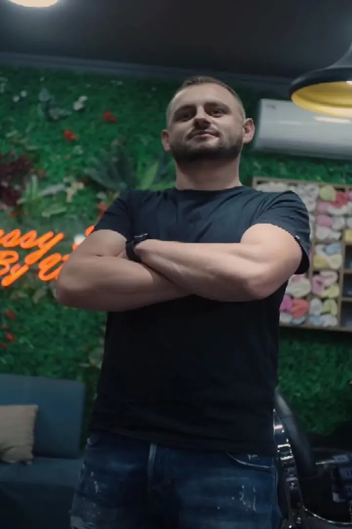A Visi barber, arms crossed, in front of the shop's green wall